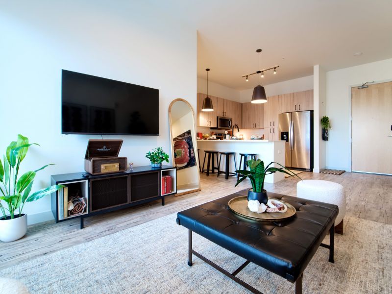 Lumen on Ninth Student Living Apartments Modern living room with a TV, plants, kitchen, and a black ottoman in a bright, open space.